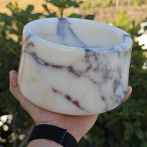 Könnte beinhalten: Eine weiße Marmorschale mit grauer und brauner Maserung. Die zylindrische Schale hat einen abgerundeten Rand und wird in einer Hand gehalten. Der Hintergrund ist verschwommen und zeigt grünes Laub.