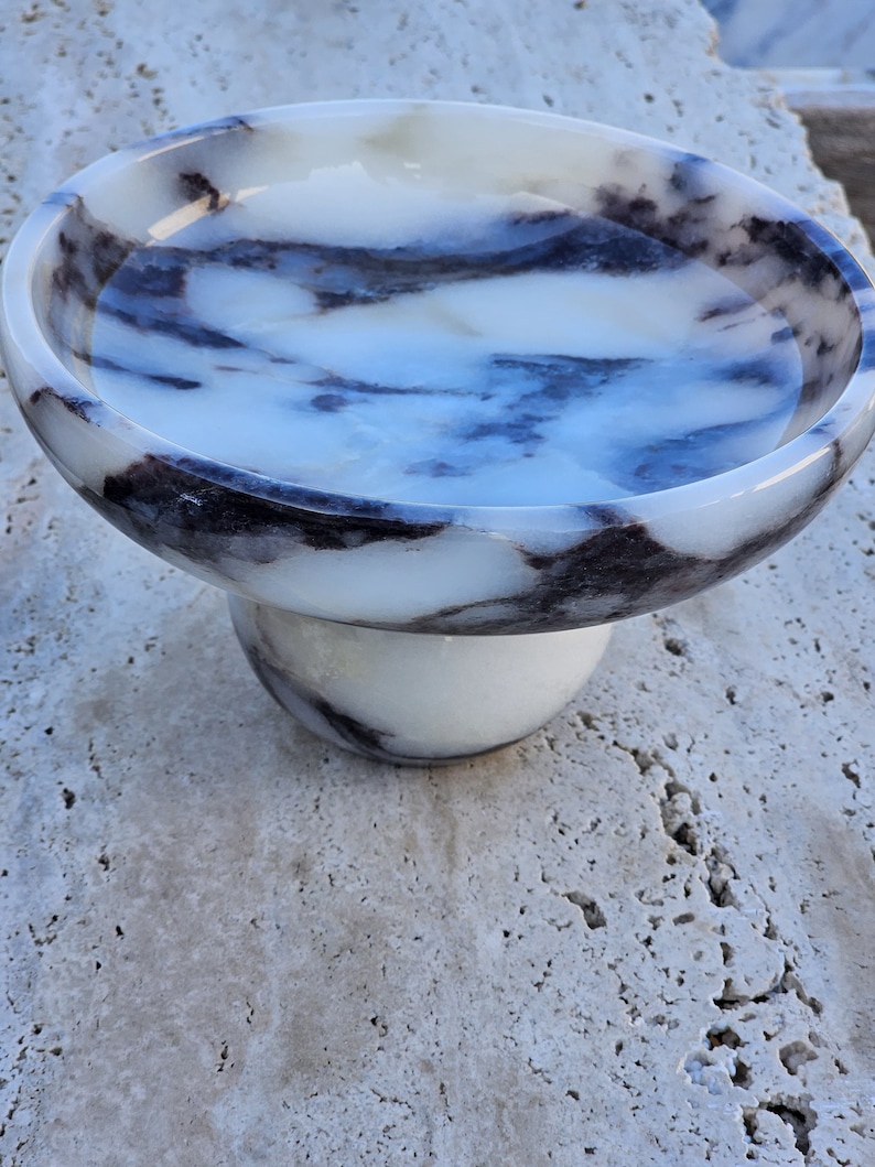 May include: A white marble bowl with black and gray veining, sitting on a white marble pedestal. The bowl is round and has a smooth surface.