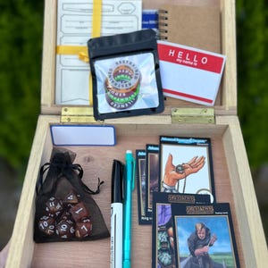 May include: A wooden box containing various gaming items. Inside are dice, trading cards, pens, an eraser, and tokens. A name tag reads "HELLO my name is". The tokens display the words "RESTRAINED, REDUT, SILENCE".