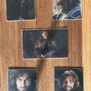 Puede incluir: Cinco tarjetas rectangulares en fundas transparentes, cada una con un retrato de un hombre con pelo largo. El hombre aparece con diferentes atuendos y expresiones. Las tarjetas est&aacute;n dispuestas sobre una superficie de madera.