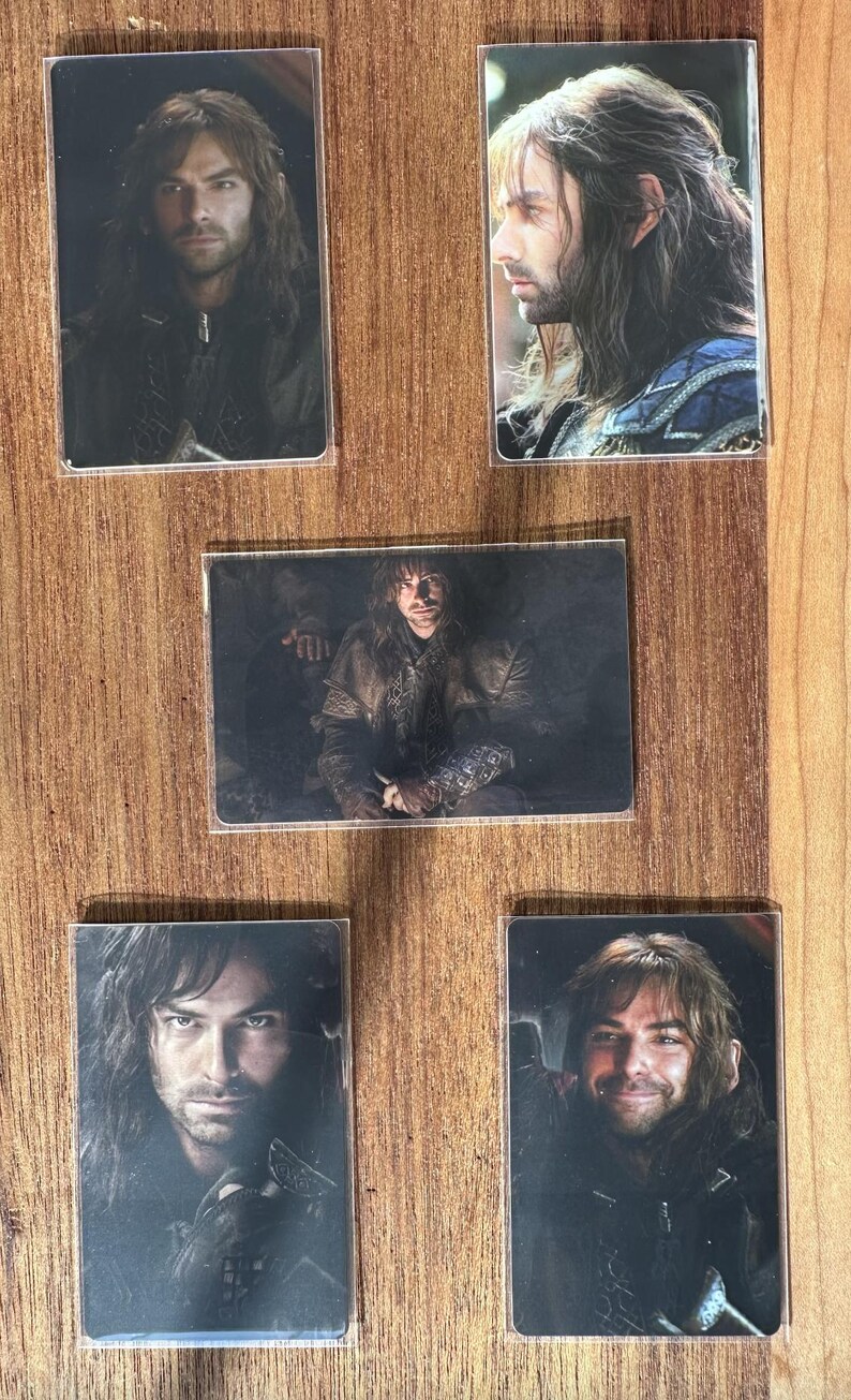 Puede incluir: Cinco tarjetas rectangulares en fundas transparentes, cada una con un retrato de un hombre con pelo largo. El hombre aparece con diferentes atuendos y expresiones. Las tarjetas est&aacute;n dispuestas sobre una superficie de madera.