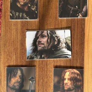 Puede incluir: Colecci&oacute;n de cinco tarjetas rectangulares con im&aacute;genes de un hombre con cabello largo y oscuro y barba. Las tarjetas est&aacute;n dispuestas sobre un fondo de madera. El hombre lleva varios atuendos, y algunas tarjetas lo muestran sosteniendo una espada.