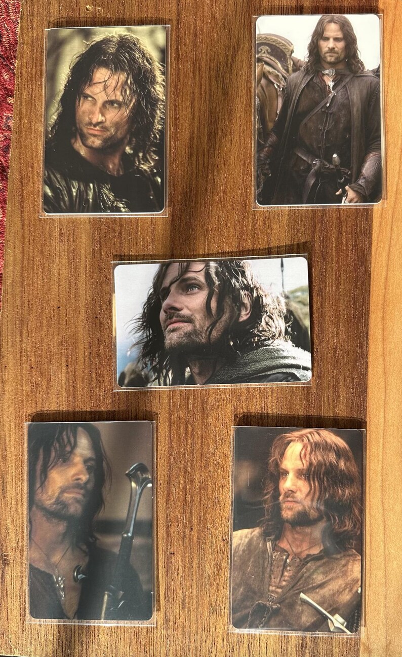 Puede incluir: Colecci&oacute;n de cinco tarjetas rectangulares con im&aacute;genes de un hombre con cabello largo y oscuro y barba. Las tarjetas est&aacute;n dispuestas sobre un fondo de madera. El hombre lleva varios atuendos, y algunas tarjetas lo muestran sosteniendo una espada.