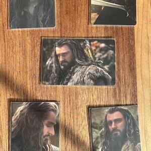 Puede incluir: Conjunto de cinco cromos que muestran a un hombre con pelo largo y ondulado y barba. Los cromos muestran diferentes poses, algunas con una espada o ropa con forro de piel. Los cromos se presentan sobre una superficie de madera.