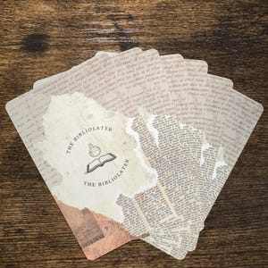 Puede incluir: Un abanico de tarjetas de estilo vintage con un efecto de papel rasgado. Las tarjetas presentan texto impreso y las palabras "THE BIBLIOLATER" con un logotipo de un libro abierto y un frasco. Las tarjetas est&aacute;n dispuestas sobre una superficie de madera.