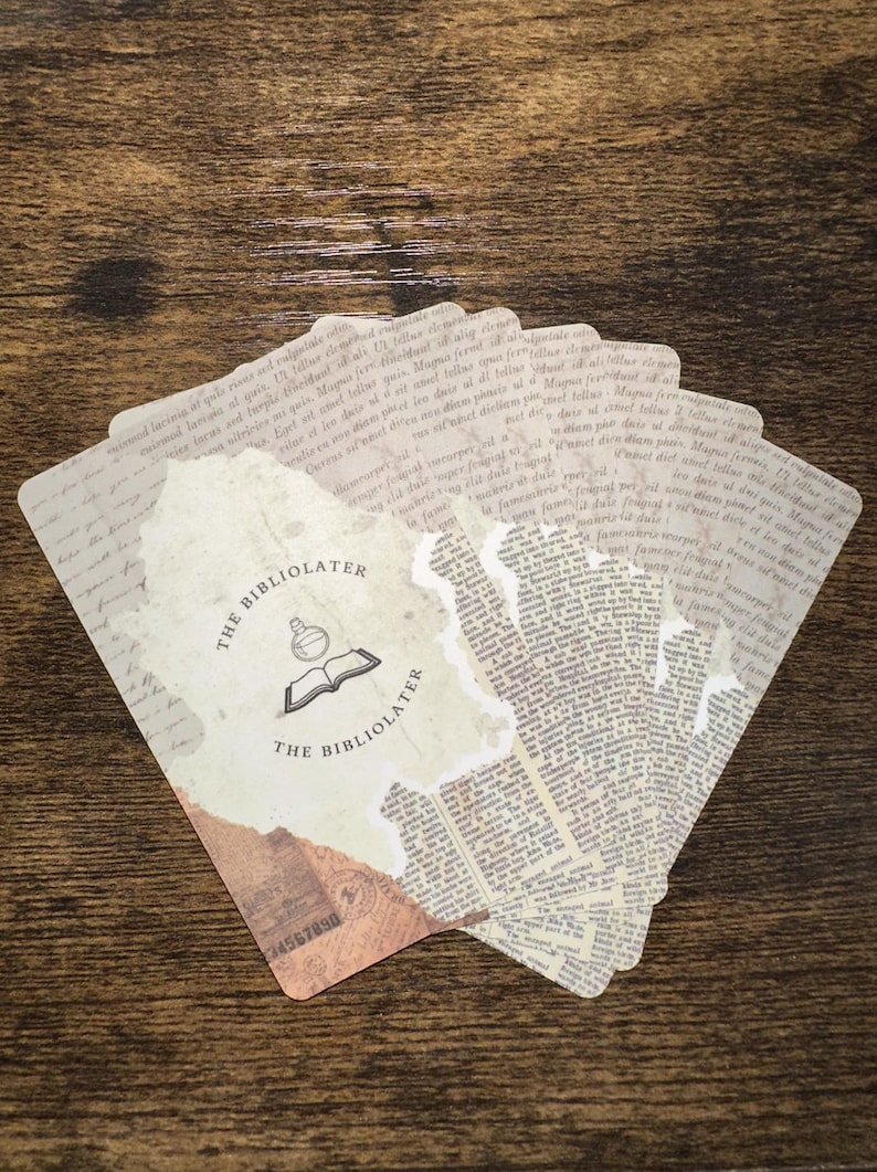 Puede incluir: Un abanico de tarjetas de estilo vintage con un efecto de papel rasgado. Las tarjetas presentan texto impreso y las palabras "THE BIBLIOLATER" con un logotipo de un libro abierto y un frasco. Las tarjetas est&aacute;n dispuestas sobre una superficie de madera.