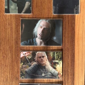 Puede incluir: Colecci&oacute;n de cinco cromos brillantes con un personaje de pelo largo y blanco. El personaje se representa en varias poses, algunas sosteniendo una espada y vistiendo ropa oscura. Las cartas se exhiben sobre una superficie de madera.