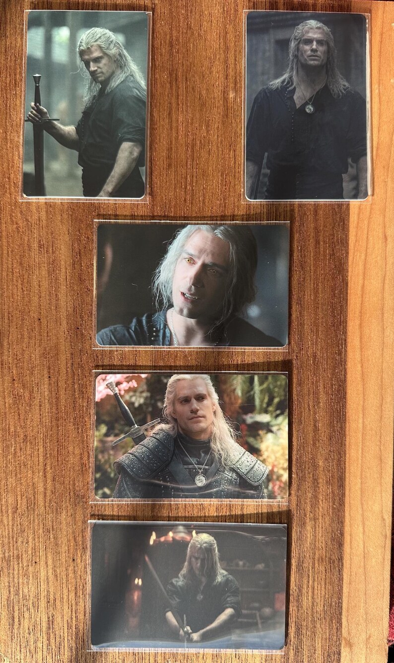 Puede incluir: Colecci&oacute;n de cinco cromos brillantes con un personaje de pelo largo y blanco. El personaje se representa en varias poses, algunas sosteniendo una espada y vistiendo ropa oscura. Las cartas se exhiben sobre una superficie de madera.