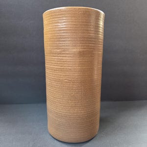 May include: A cylindrical brown ceramic vase with horizontal ribbed texture. The vase has a slightly flared rim and a smooth, unglazed finish. The vase is a neutral color and could be used for flowers or as a decorative item.