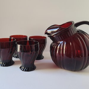 May include: A vintage ruby red glass pitcher and four matching glasses. The pitcher has a ribbed design and a handle. The glasses have a similar ribbed design and a flared base. The set is likely for serving beverages.