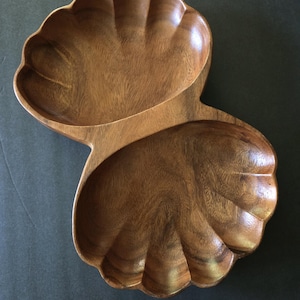 May include: A wooden serving dish with a shell-like design. The dish is made of a light brown wood and has two connected compartments. The edges of the dish are scalloped, and the wood grain is visible.