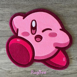 Puede incluir: Alfombra de felpa rosa y roja con forma del personaje de videojuego Kirby. Kirby est&aacute; gui&ntilde;ando un ojo y sonriendo con la boca abierta. La alfombra tiene un fondo rosa y detalles rojos.