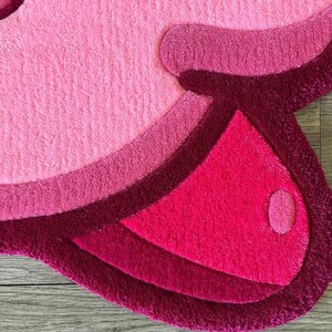 Puede incluir: Una alfombra peluda rosa y roja con un dise&ntilde;o ondulado. La alfombra tiene forma de nube y una textura suave y mullida.
