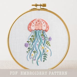 May include: Embroidered jellyfish design in a gold hoop. The jellyfish is pink and white with blue tentacles, surrounded by embroidered flowers and greenery. The text "PDF EMBROIDERY PATTERN" is at the bottom.