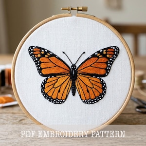 May include: Embroidered monarch butterfly on white fabric stretched in a wooden hoop. The butterfly features orange and black wings with white spots. The text "PDF EMBROIDERY PATTERN" is at the bottom.