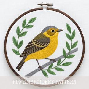 May include: An embroidery hoop containing a white fabric panel with a hand-stitched design of a yellow and brown bird perched on a gray branch, framed by two green leafy sprigs. The text PDF EMBROIDERY PATTERN is printed at the bottom.