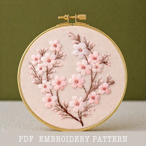 May include: A round embroidery hoop featuring a floral design. The design showcases pink and white flowers with brown stems, stitched on a light beige fabric. The hoop is gold-colored, and the text "PDF EMBROIDERY PATTERN" is visible at the bottom.
