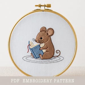 May include: An embroidery hoop featuring a brown mouse reading a blue book. The mouse is sitting on a gray stitched circle. The embroidery is set in a gold hoop. The text "PDF EMBROIDERY PATTERN" is at the bottom.