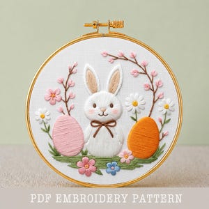 May include: Embroidered Easter design in a gold hoop. A white bunny with a brown bow tie is centered between a pink and orange egg. Pink flowers and green grass surround the bunny. The text "PDF EMBROIDERY PATTERN" is at the bottom.