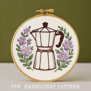 May include: Embroidered coffee maker design in a gold hoop. The brown coffee maker is surrounded by purple flowers and green leaves. The text "PDF EMBROIDERY PATTERN" is at the bottom of the image.