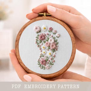 Puede incluir: Un bastidor de bordado circular de madera que contiene tela blanca con un diseño floral en forma de huella de bebé. El patrón presenta rosas rosas bordadas, pequeñas margaritas blancas y hojas verdes. El texto PDF EMBROIDERY PATTERN es visible.