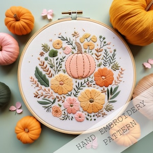 以下が含まれることがあります： ピンクのかぼちゃがオレンジ、イエロー、ピンクの花と緑の葉に囲まれた刺繍のフープアート。フープは緑色の背景に他のカボチャと花が置かれています。画像の右下隅には「HAND EMBROIDERY PATTERN」というテキストがあります。