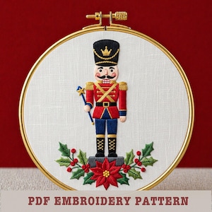 May include: Embroidered Christmas decoration in a gold hoop, featuring a nutcracker soldier. The soldier has a red jacket, blue trousers, and a black hat. Holly and a poinsettia are at the base. Text at the bottom reads "PDF EMBROIDERY PATTERN".