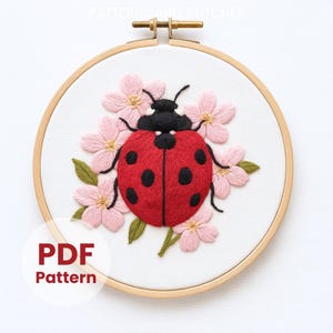 Puede incluir: Diseño de mariquita bordada sobre tela blanca, en un bastidor de bordado de madera. La mariquita es roja con lunares negros, rodeada de flores rosas y hojas verdes. El texto "PDF Pattern" es visible.