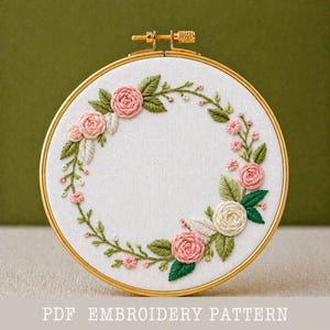 Könnte beinhalten: Ein Stickrahmen mit floralem Design. Das Design zeigt rosa und weiße Rosen, grüne Blätter und kleine rosa Blüten, die in einem Kranz angeordnet sind. Der Text "PDF EMBROIDERY PATTERN" befindet sich unten.