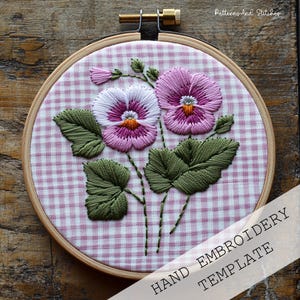 Könnte beinhalten: Handstickvorlage mit zwei rosa und weißen Stiefmütterchen mit grünen Blättern auf einem rosa und weißen Gingham-Stoff. Die Vorlage befindet sich in einem hölzernen Stickrahmen. Der Text "HAND EMBROIDERY TEMPLATE" befindet sich am unteren Rand des Bildes.