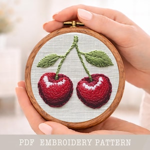 Puede incluir: Un bastidor de bordado cosido a mano con dos cerezas rojas con detalles blancos, hojas y tallos verdes. El bordado está sobre una tela clara, enmarcado por un aro de madera. El texto "PDF EMBROIDERY PATTERN" está en la parte inferior.