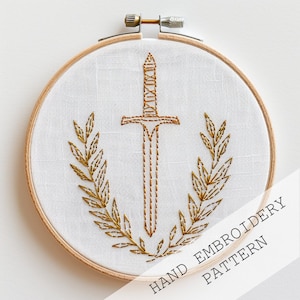 May include: Gold embroidery pattern of a sword with a laurel wreath on a white fabric in a wooden embroidery hoop. The text "HAND EMBROIDERY PATTERN" is printed on the bottom right corner of the image.