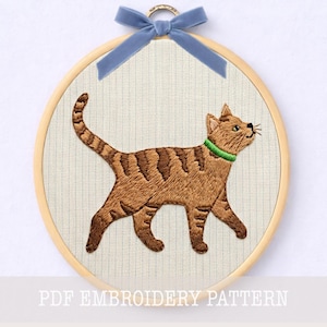 May include: A circular wooden embroidery hoop containing a fabric panel with a stitched brown tabby cat. The cat has dark brown stripes, a bright green collar, and is walking towards the right. A blue velvet ribbon is tied at the top of the hoop.