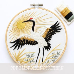 May include: A circular wooden embroidery hoop containing a white fabric panel with a hand-stitched crane in flight. The crane features white, black, and gold thread with a red crown. A golden sun and stylized grass surround the bird. A thread card is included.