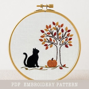 Può includere: Ricamo in un cerchio dorato. Raffigura un gatto nero, un albero con foglie autunnali nei toni del rosso, arancione e giallo e una zucca arancione. Lo sfondo è in tessuto bianco. Il testo in basso recita "PDF EMBROIDERY PATTERN".