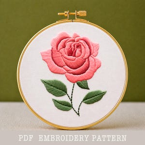 May include: A detailed embroidery of a pink rose with green leaves and stem, set in a gold-toned hoop. The text "PDF EMBROIDERY PATTERN" is at the bottom. The rose is the focal point, showcasing intricate stitching on white fabric.