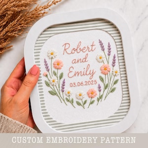 May include: White framed embroidery with a floral design. The embroidery features the names "Robert and Emily" and the date "03.06.2025" in pink thread. The frame has a striped background and the text "CUSTOM EMBROIDERY PATTERN".