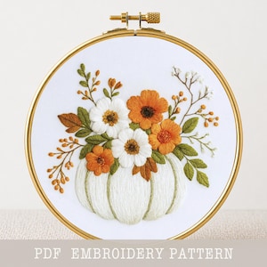 May include: A round gold embroidery hoop containing white fabric with a hand-stitched design of a white pumpkin topped with a floral arrangement of white and orange flowers, green leaves, and small orange berries. The text PDF EMBROIDERY PATTERN is visible.