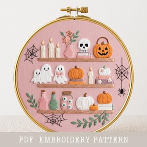 Pode incluir: Um bastidor de bordado circular com tecido rosa contendo prateleiras bordadas exibindo três velas, um vaso de flores, uma caveira, abóboras, fantasmas, frascos de poções e globos oculares. Teias de aranha pretas e uma aranha estão bordadas.