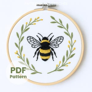 以下が含まれることがあります： 木製のフープに刺繍された蜂のデザイン。 蜂は黒と黄色で、緑の葉と黄色の花に囲まれています。 「PDF Pattern」というテキストが下部の緑色の円の中にあります。 上部には「PATTERNS AND STITCHES」という文字があります。