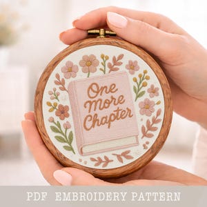 Peut inclure: Un cercle à broder rond avec un motif floral et le texte "One More Chapter". La broderie présente un livre rose entouré de fleurs roses et pêche et de feuilles vertes. Le cercle est tenu dans les mains d'une personne.
