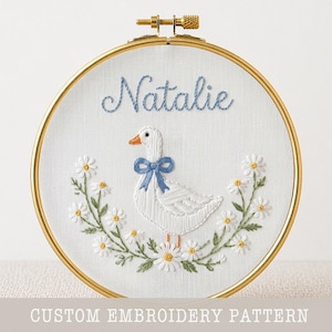 May include: Embroidered hoop art with a white goose wearing a blue bow, encircled by a floral wreath. The name "Natalie" is stitched in blue above the goose. The gold hoop holds the white fabric. Text at the bottom reads "CUSTOM EMBROIDERY PATTERN".