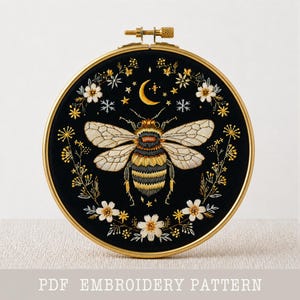 Könnte beinhalten: Ein schwarzer Stickrahmen mit einem detaillierten Bienenmuster, einem Mond und Sternen. Die Biene hat hellbraune Flügel und einen gelb-schwarz gestreiften Körper. Das Design ist von weißen und gelben floralen Akzenten umgeben. Der Text "PDF EMBROIDERY PATTERN" steht unten.