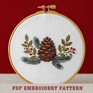 May include: Embroidered pinecone and foliage design in a gold hoop. The design features a detailed brown pinecone, green leaves, and red berries on a white fabric. The text "PDF EMBROIDERY PATTERN" is at the bottom.