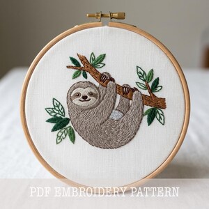 Pode incluir: Bordado de um bicho-preguiça pendurado em um galho, rodeado por folhas verdes, dentro de um bastidor de madeira. A preguiça é cinza e marrom, com um rosto sorridente. O texto na parte inferior diz "PDF EMBROIDERY PATTERN".