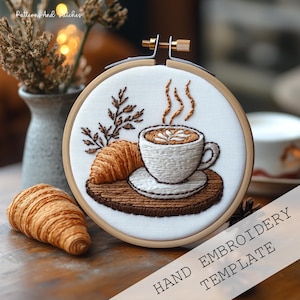 Puede incluir: Plantilla de bordado a mano con una taza de café con arte latte, un cruasán y hojas. El diseño está bordado sobre tela blanca dentro de un bastidor de madera. El texto "HAND EMBROIDERY TEMPLATE" es visible.