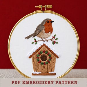 May include: Embroidered robin perched on a branch above a birdhouse, set in a gold hoop. The bird has a red breast and brown wings. The birdhouse is brown with a wreath and snow detail. Text at the bottom reads "PDF EMBROIDERY PATTERN".