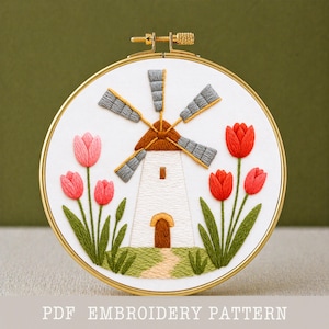 Pode incluir: Um bastidor de bordado circular contendo tecido branco com um desenho bordado à mão de um moinho de vento branco com telhado castanho e pás cinzentas, rodeado por tulipas vermelhas e cor-de-rosa com caules e folhas verdes e um caminho.