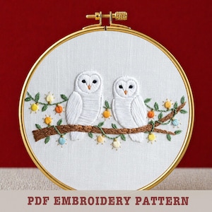 May include: Embroidered hoop art featuring two white owls perched on a brown branch with green leaves and colorful accents. The owls have black eyes and are set against a white background within a gold hoop. Text at the bottom reads "PDF EMBROIDERY PATTERN".