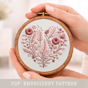 Könnte beinhalten: Ein herzförmiges Stickdesign mit rosa Blumen und Blättern auf weißem Grund, in einem Holzreifen. Der Text "PDF EMBROIDERY PATTERN" befindet sich am unteren Rand des Bildes.